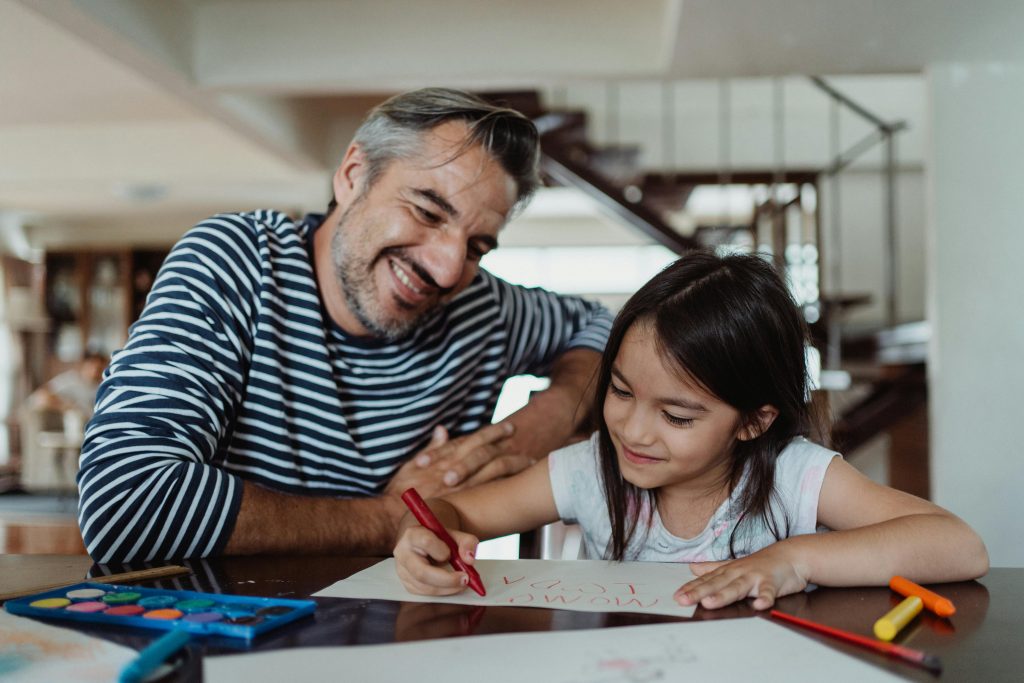 girl writing on paper using red crayon with adult male at table